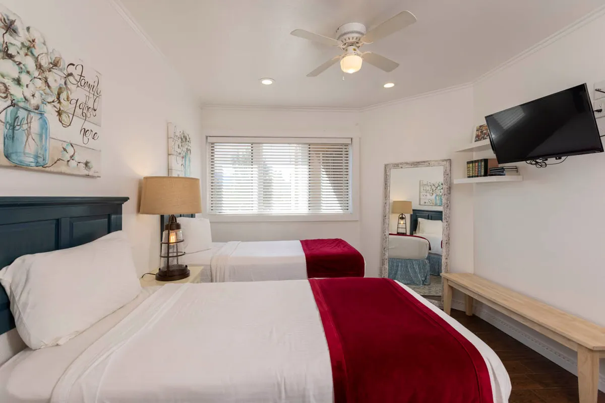 A cozy bedroom with two beds dressed in crisp white linens and red accents, flanked by bedside tables and lamps. Decorative wall art adds character, while a mounted television and full-length mirror provide modern convenience and practicality.