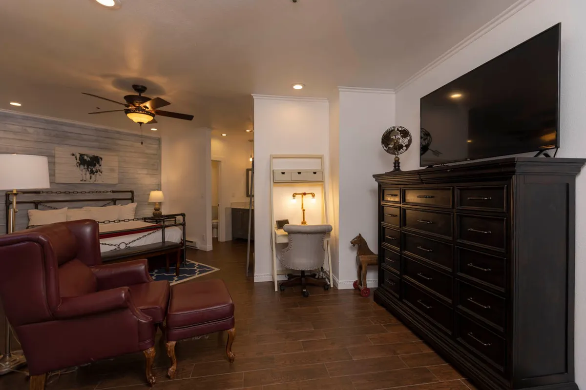 A cozy multi-purpose bedroom with a bed, dressing table, armchair, television, and natural lighting. The space feels inviting for both relaxation and remote work, with surfaces conducive to working or unwinding.