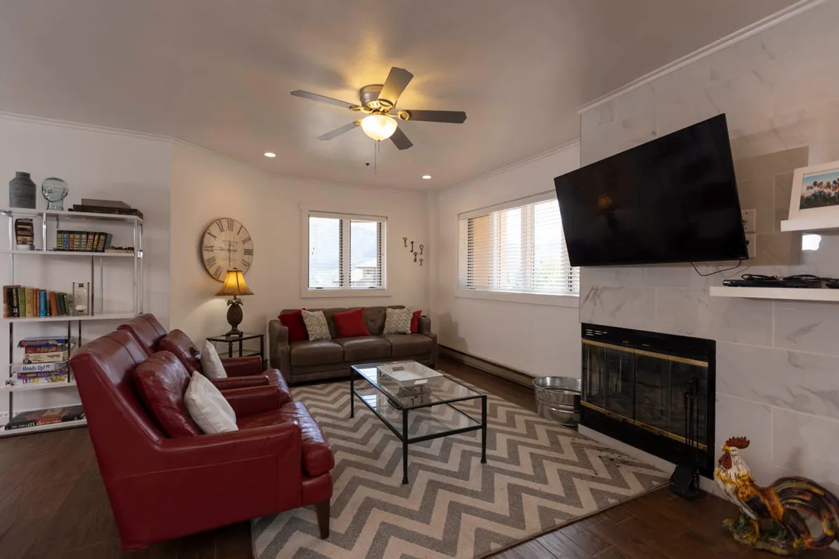 A cozy condo living room featuring a glowing wood-burning fireplace with a TV mounted above, surrounded by a plush red leather sofa set, a glass coffee table, and a bookshelf stocked with books and games. The space invites relaxation, socializing, and unwinding after mountain adventures.