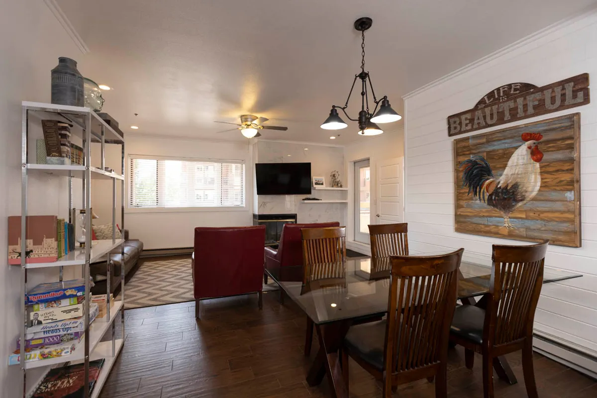 A cheerful, multi-purpose gathering space with a wood-burning fireplace beneath a wall-mounted TV. The glass dining table with wooden chairs offers a spot for shared meals or games, while rustic decor—including a prominent rooster artwork and 'Life is Beautiful' sign—brings positivity and mountain charm to the space. Books and games on the metal shelving unit invite group activities.