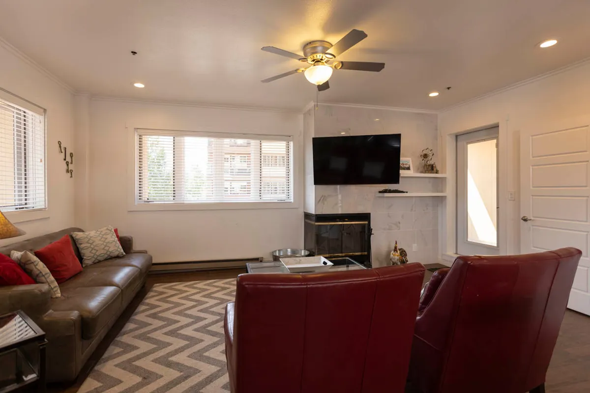 The interior scene centers on a cozy living area with a wood-burning fireplace, two red leather chairs, a large brown leather sofa, and a chevron-patterned rug. The inviting arrangement suggests a space for warming up, relaxing, and enjoying quiet moments after outdoor pursuits.