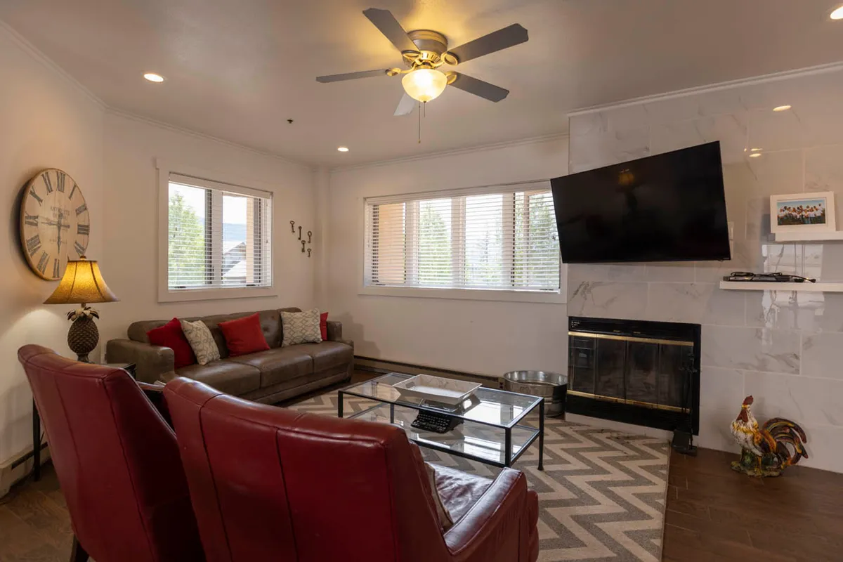 A warm and welcoming living room anchored by a wood-burning fireplace with a TV mounted above. The brown leather couch with red and patterned pillows, two red leather armchairs, and a glass coffee table create an inviting space for relaxation and social gatherings. Large windows offer glimpses of tranquil mountain views, amplifying the cozy lodge atmosphere.