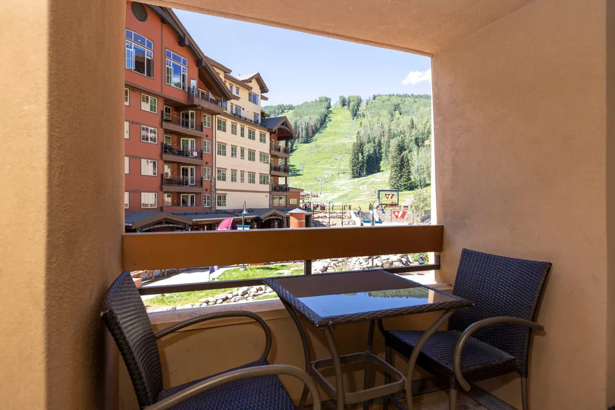 This image shows a balcony seating area, complete with a table and chairs, overlooking green ski slopes, forested mountains, and an adjacent lodge building. The setting suggests a serene spot for morning coffee, meals, or unwinding while immersed in nature.