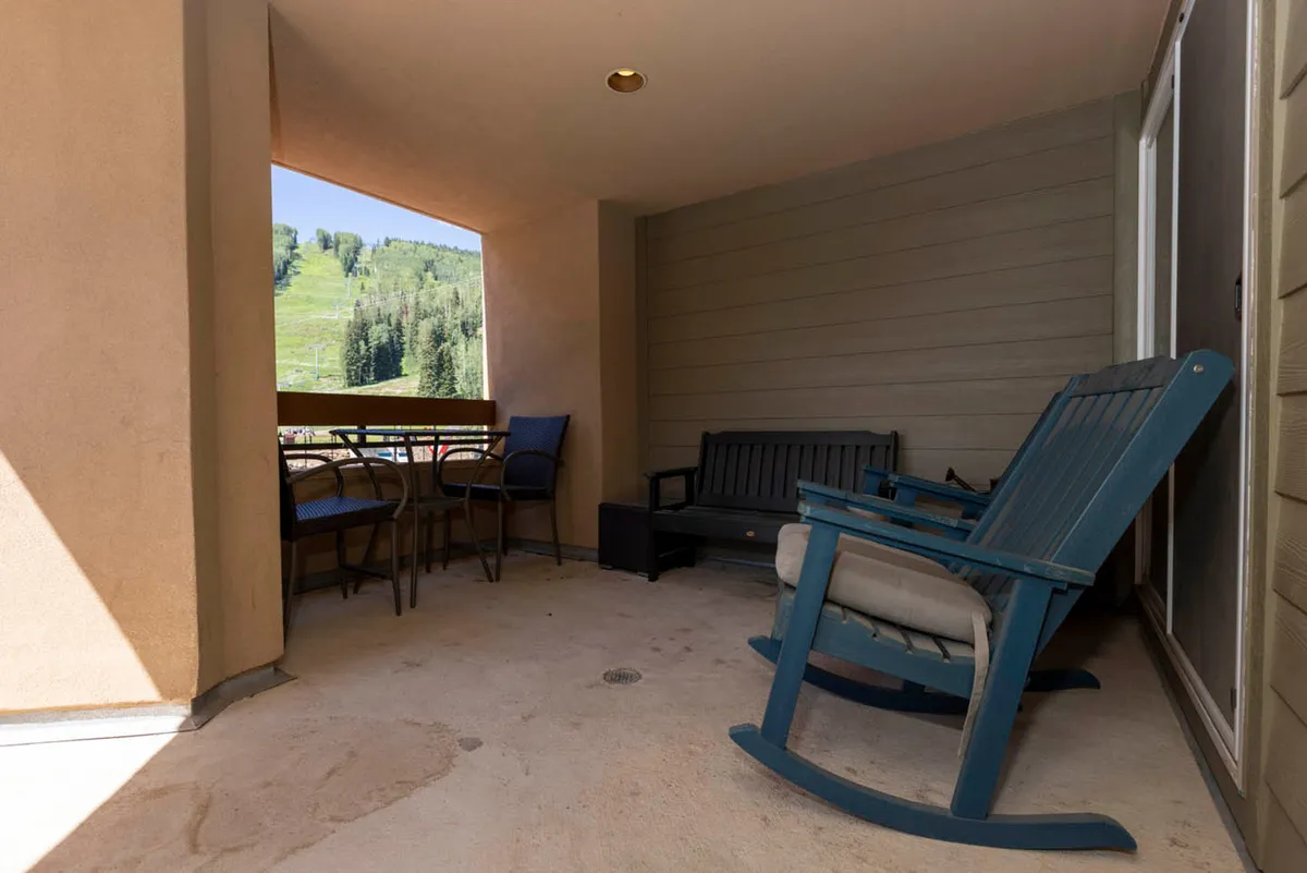 An outdoor balcony featuring blue wooden rocking chairs with cushions, a table with four chairs, a black bench along the wall, and a scenic view of a green mountain slope through the balcony opening. The arrangement invites socializing and relaxation while soaking in mountain vistas.