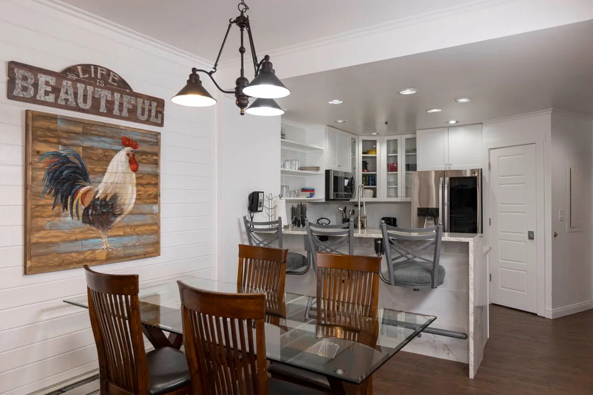 A bright and uplifting kitchen and dining area with a glass-top dining table surrounded by wooden chairs. The kitchen island with bar stools offers casual seating for meal prep or breakfast. Uplifting decor, including large rooster wall art and a 'Life is Beautiful' sign, enhances the warm, communal vibe.
