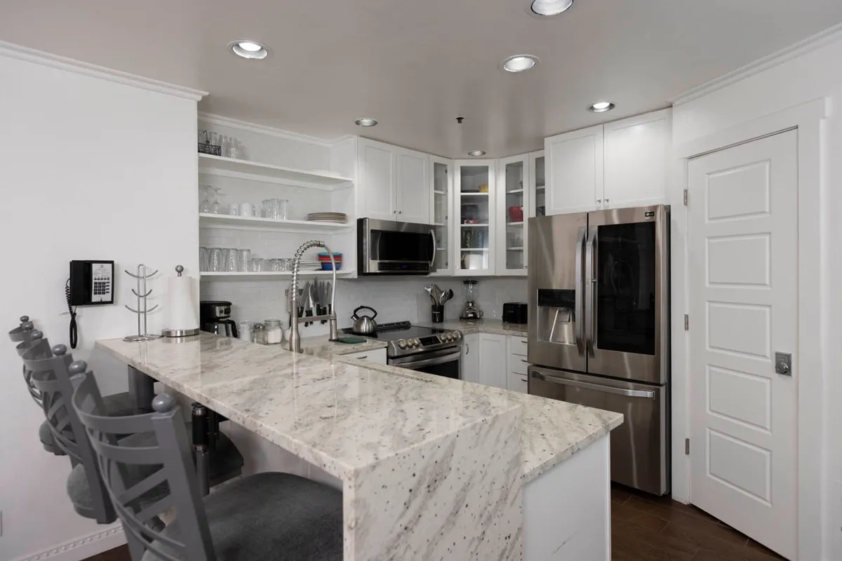 An inviting interior space featuring a marble kitchen island with bar stools, stainless steel appliances, and open shelving stocked with glassware. The setting is warm, functional, and ideal for socializing or enjoying a quick meal before heading out.