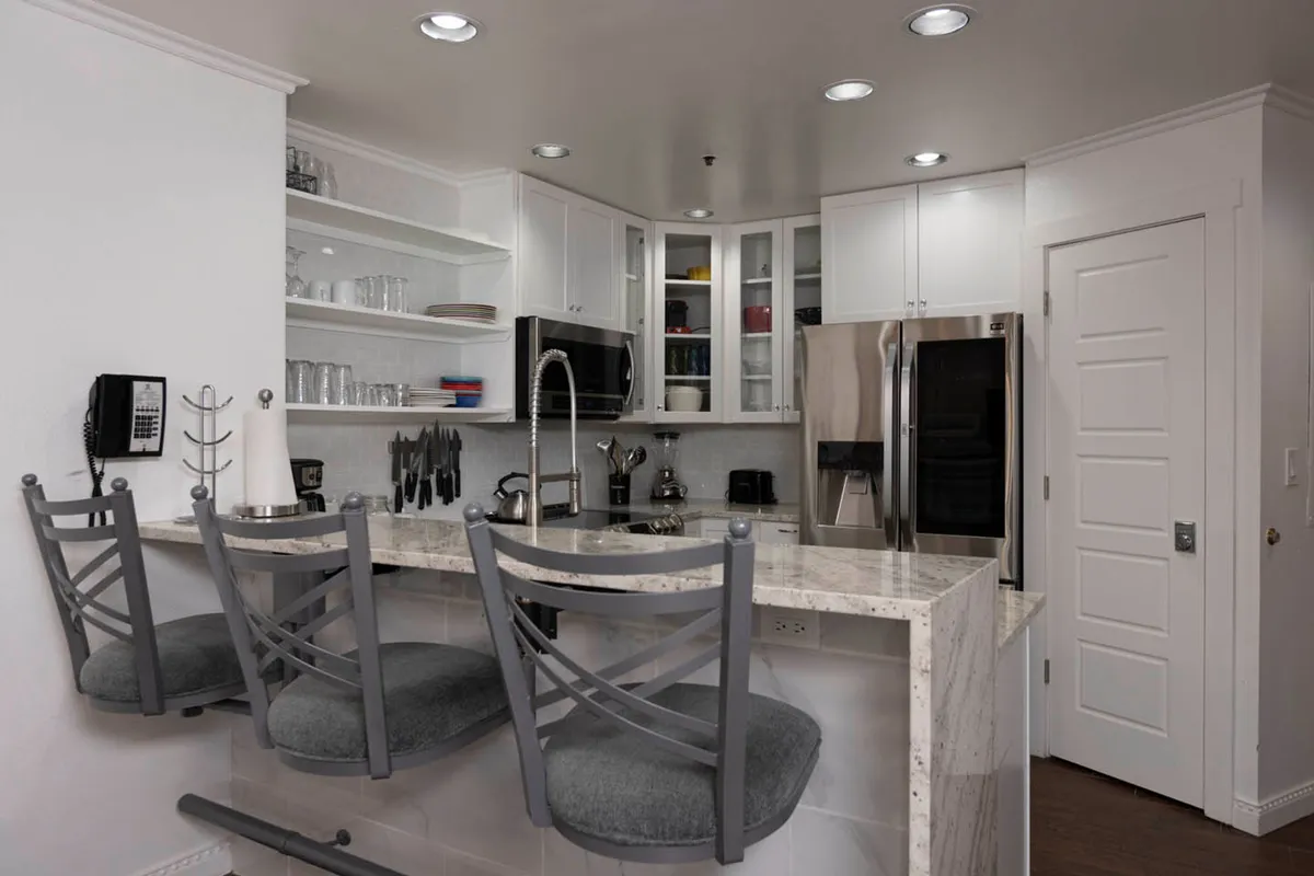 A bright, inviting kitchen with a marble island that doubles as seating, complemented by a stainless steel refrigerator, built-in microwave, and open shelving with glassware. The space is designed for meal preparation, casual dining, and social gatherings.