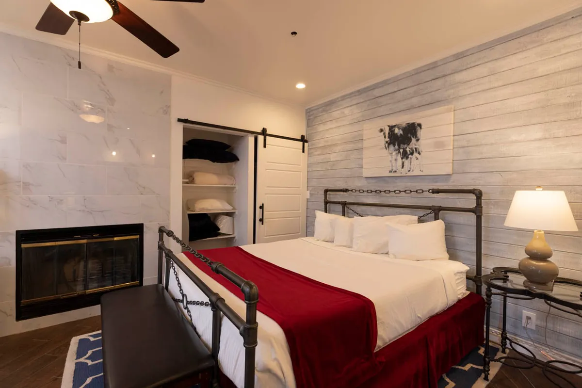 A warmly lit bedroom featuring a large bed with soft white linens and a red throw blanket sits near a wood-burning fireplace with a marble surround. The decor includes a barn-style sliding door revealing a linen closet and a decorative wall art of a cow above the bed, all contributing to a cozy, restful mountain ambiance.