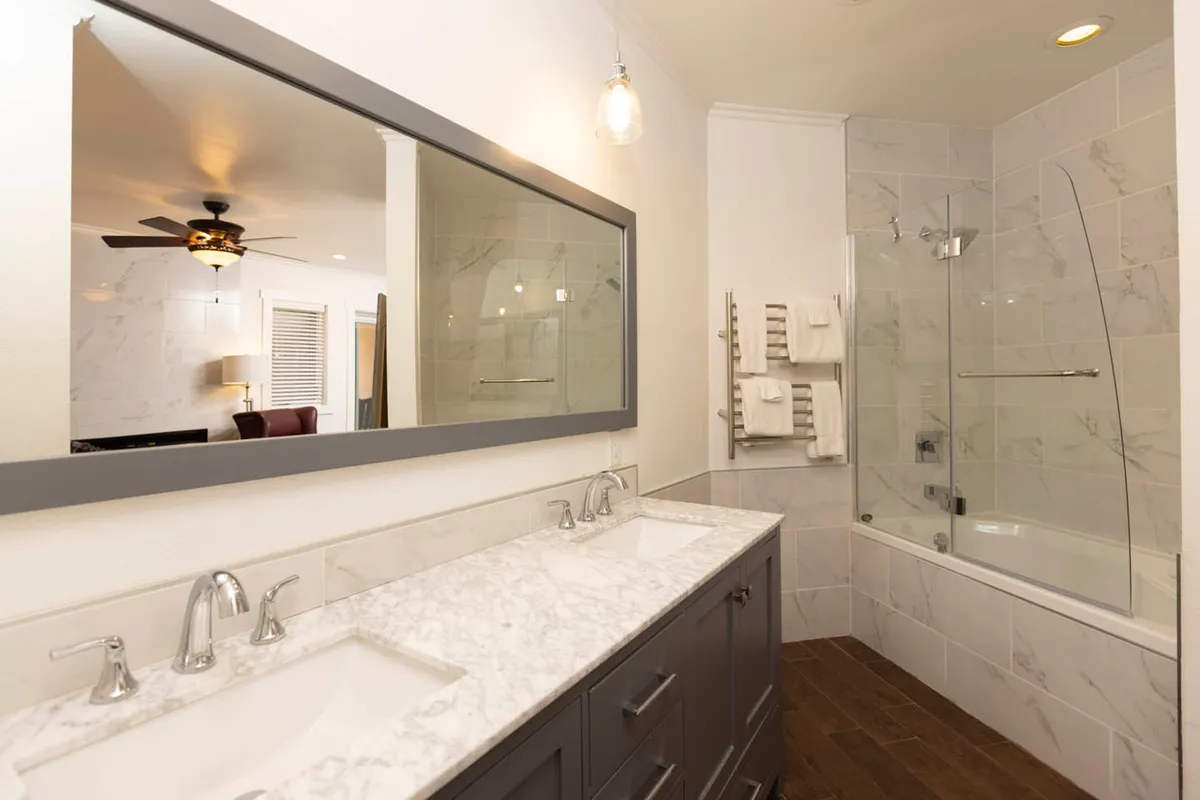 A spa-inspired bathroom with a double marble sink vanity and a large mirror, accompanied by a glass-enclosed bathtub/shower area and a heated towel rack with neatly arranged towels. The space feels bright, clean, and luxurious, ideal for unwinding.