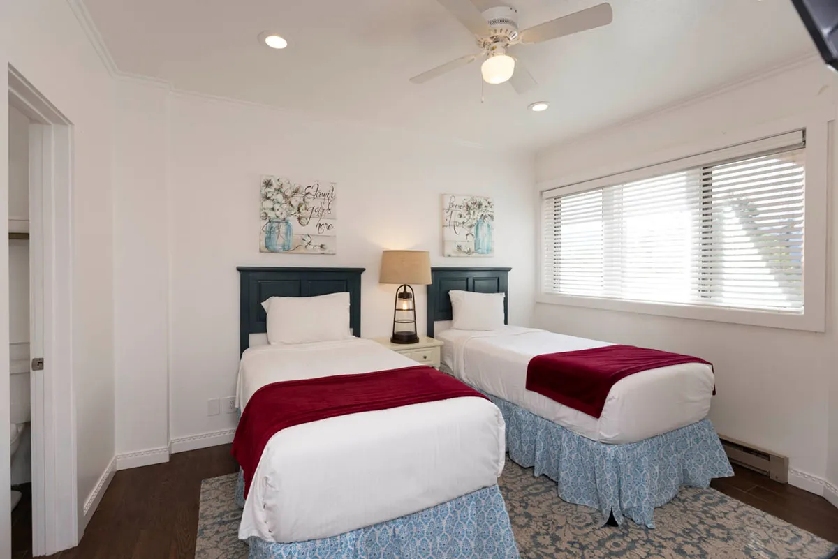 A tranquil bedroom with two twin beds, each topped with white linens and red throws. A nightstand with a lamp sits between the beds, and a large window with white blinds allows natural light to filter in. A ceiling fan with a light fixture above completes the serene scene.