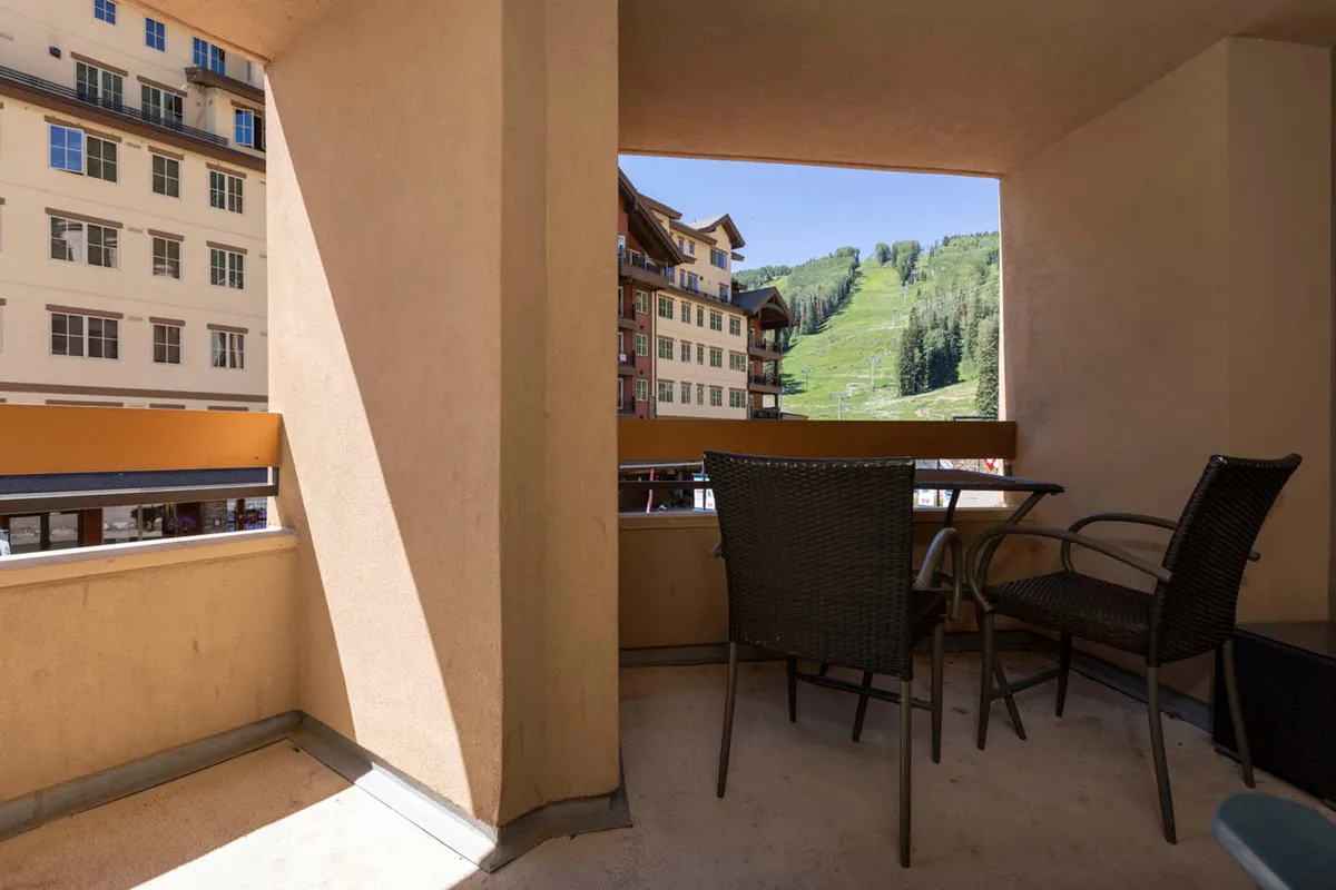 This image presents a private balcony furnished with two black wicker chairs and a small round glass table, overlooking a stunning mountain vista and the adjacent Purgatory Village buildings below. The balcony setting invites occupants to relax, enjoy the fresh air, and take in the tranquil natural surroundings while being close to village amenities.