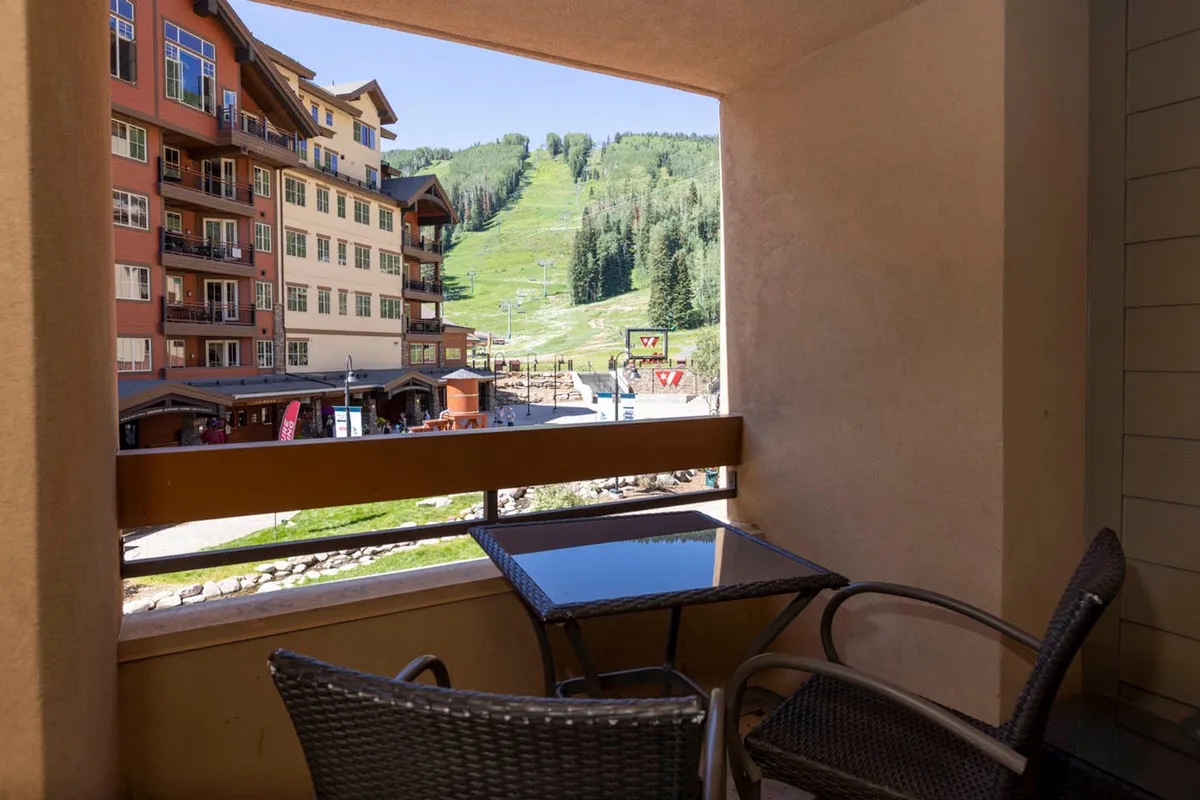 A peaceful balcony set with a table and chairs overlooks the ski slopes, chairlift, surrounding green mountains, and adjacent resort buildings. The setting is ideal for relaxing while observing mountain activities or planning the next adventure.