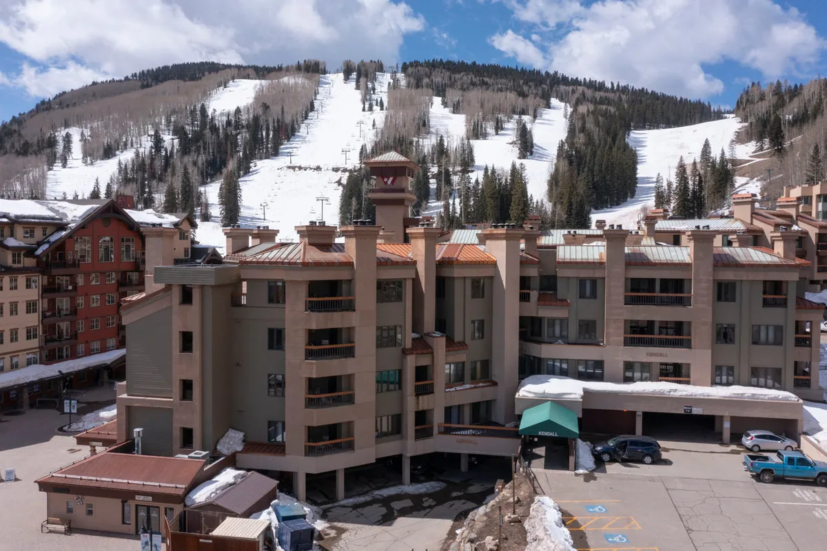 An exterior view highlighting the multi-story lodge building with balconies overlooking snowy ski slopes, a parking area with vehicles in the foreground, and a bright blue sky. The scene emphasizes easy access to both mountain adventures and practical amenities like garage parking.