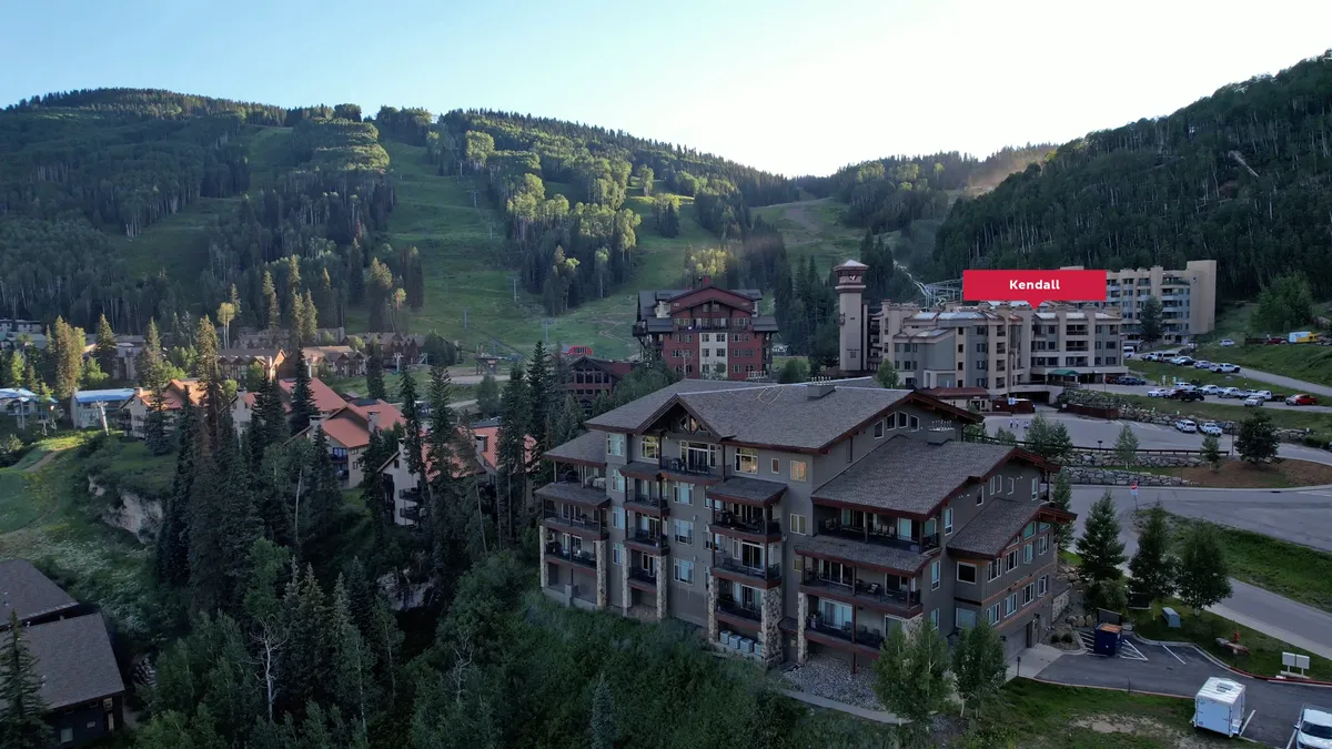 A wide exterior shot of Kendall Mountain Lodge, featuring balconies overlooking pine forests and mountain slopes. The Village Plaza and nearby buildings can be seen in the foreground, while ski runs and the surrounding landscape emphasize the proximity to outdoor activities and resort amenities.