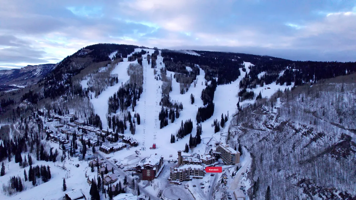 A vibrant exterior scene showing the Kendall Mountain Lodge complex with visible ski slopes, ski lifts, and the adjacent Village Plaza area. The image captures snow-covered slopes, the bustling plaza with nearby shops and amenities, and the inviting lodge buildings nestled at the heart of the village.
