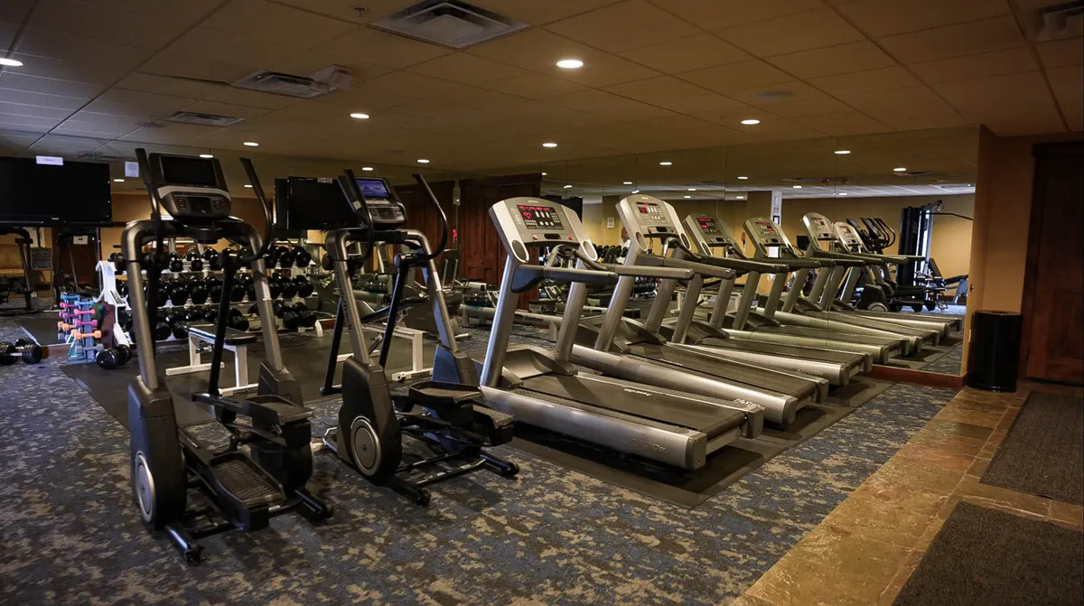 A well-equipped fitness center with a row of treadmills, elliptical machines, a dumbbell rack, and exercise mats. The space is bright, modern, and inviting, designed for both cardio and strength workouts.