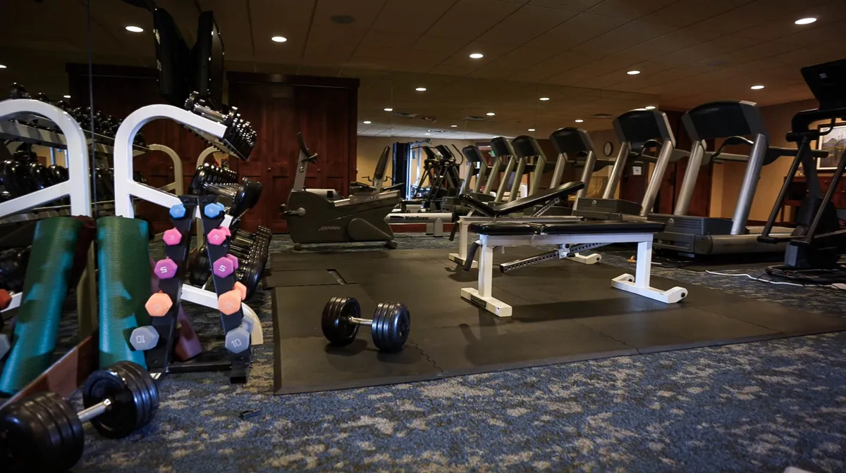 The image showcases a well-equipped fitness center, featuring a dumbbell rack, weight bench, treadmills, and exercise mats. The space is bright and functional, emphasizing opportunities for wellness and daily routines during extended stays.