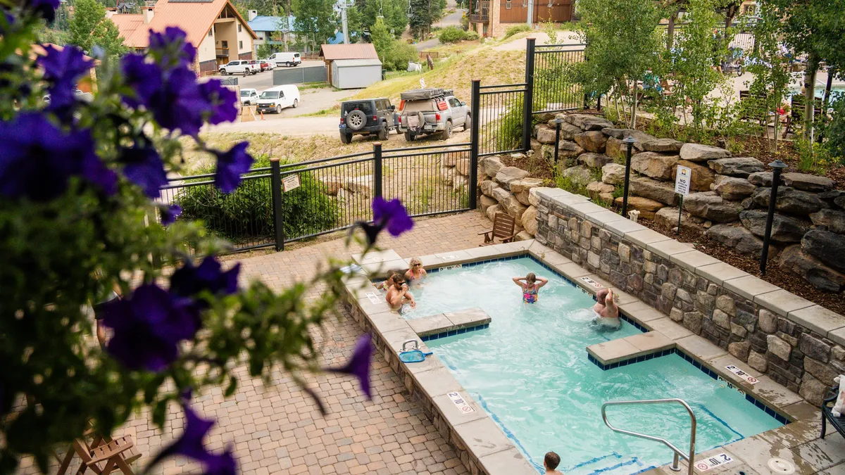 A heated outdoor swimming pool surrounded by a stone retaining wall and vibrant purple flowers, with people enjoying the warm water and socializing in a scenic mountain setting.