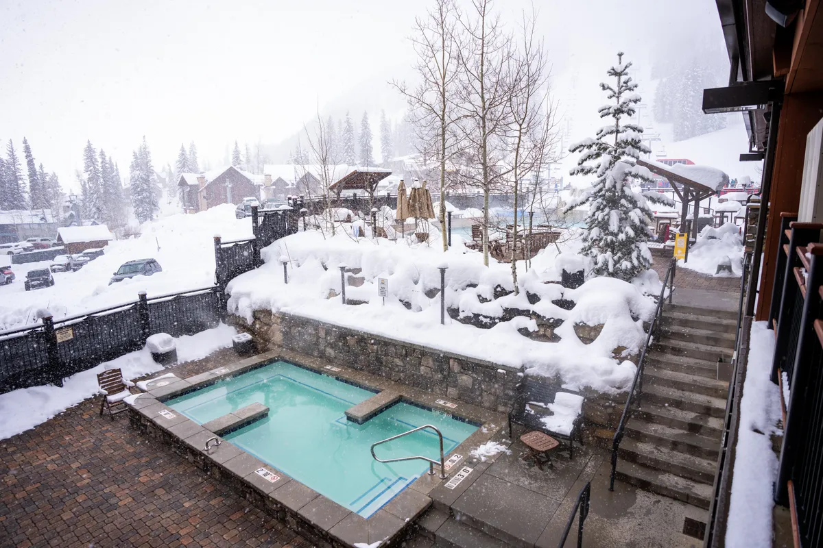A heated outdoor swimming pool with clear turquoise water bordered by snowy stone walls and steps, rustic outdoor furniture, and views of snow-dusted pine trees and a distant village. The setting is peaceful, blending outdoor winter charm with cozy comfort.
