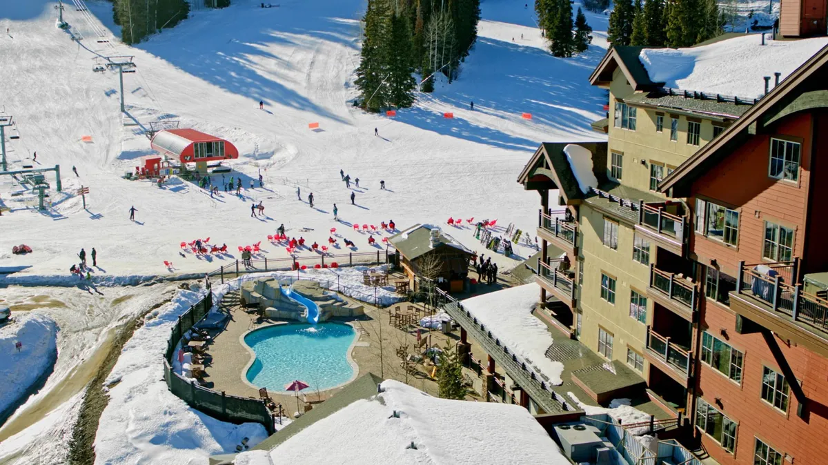 A vibrant exterior scene showing a heated outdoor swimming pool beside the lodge, with red lounge chairs and views of skiers on snowy slopes in the background. The lodge's balconies and implied rooftop hot tub add to the sense of post-adventure relaxation.
