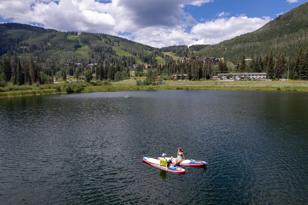 This exterior image showcases two people paddleboarding on a serene lake surrounded by mountain hillsides. The calm water, blue sky with scattered clouds, and the backdrop of scattered buildings create a sense of tranquil outdoor adventure and relaxation in a pristine alpine setting.