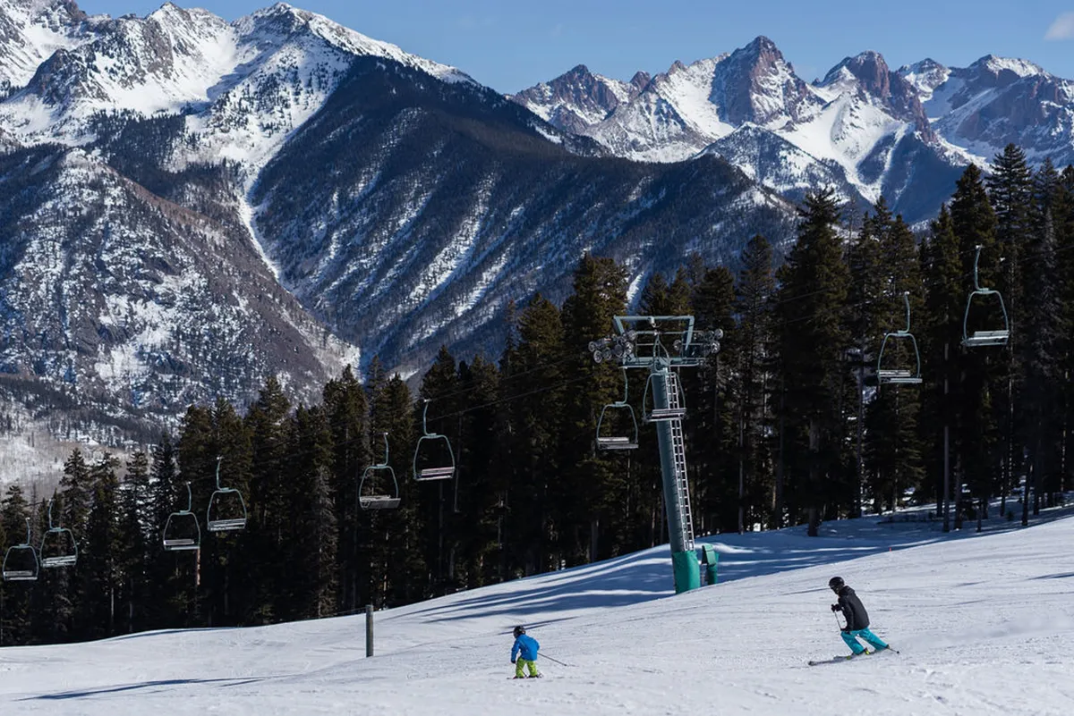 A dynamic winter scene featuring snow-covered mountain peaks, a ski chairlift in operation, skiers and snowboarders gliding down the slopes, and a dense pine forest framing the action. The image radiates excitement and the thrill of direct mountain access.
